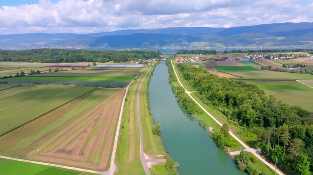 Aerial view of a vibrant canal cutting through lush green fields and rural farmland under a cloudy sky, Hagneck, Bern, Switzerland.