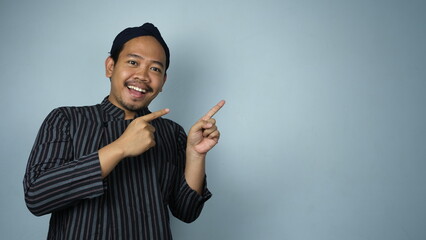 A cheerful Javanese man in traditional striped attire and cap smiling while pointing to the side,...