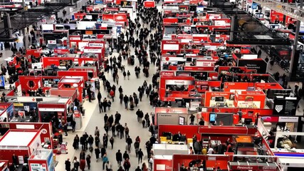 Aerial view of crowded trade show floor with numerous red-themed booths and attendees walking through wide aisles, showcasing a bustling business exhibition environment.
