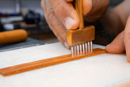 Close-up of a craftsman using a brass pricking iron on a leather strap. Precision in leatherworking: An artisan marking stitch holes on leather.
- Powered by Adobe