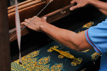 Close up of artisan hands weaving traditional fabric on a wooden loom with golden thread. Cultural handmade textile craft and craftsmanship detail."