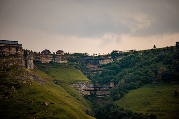 Naklejka premium A scenic view of a rocky cliff in the Caucasus region, featuring lush greenery and a small waterfall. A viewing platform is visible on the cliff edge.