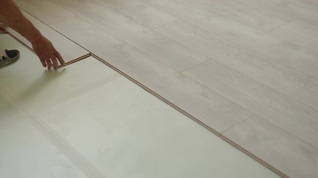 A worker carefully lays down laminate flooring in a residential space, ensuring precise placement. The light enhances the wood-like texture and color of the planks, creating a fresh atmosphere.