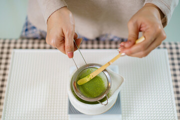 Asian woman preparing traditional Japanese matcha tea at home using bamboo tools, sifting matcha powder with care and precision.