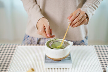 Asian woman preparing traditional Japanese matcha tea at home using bamboo tools, sifting matcha powder with care and precision.