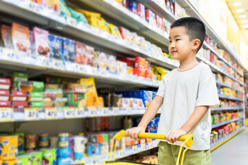A young boy is pushing a shopping cart while standing in a supermarket aisle filled with various food items.