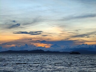 Vibrant sunset over the calm ocean, with a lone boat sailing towards the distant horizon under a sky painted with warm orange and gold twilight
