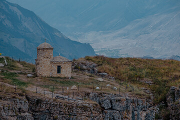 Fototapeta premium A historic stone building sits on a rocky hillside in the Caucasus Mountains. The landscape features rugged terrain and distant peaks under a cloudy sky.