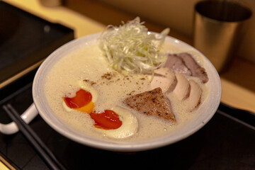 Close-up of seared meat slice being lifted with chopsticks and spoon from creamy foam ramen, soft yolk eggs and shredded leeks in broth