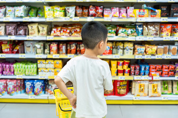 A young boy is seen from behind standing in a supermarket aisle surrounded by various snack products on the shelves.