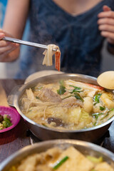 A person lifting crab stick and noodles from a steaming Taiwanese hot pot with chopsticks during a casual meal at a wooden dining table
