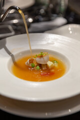 Refined scallop and vegetable soup served in a wide white bowl, hot consommé being poured at the table in a luxurious dining environment