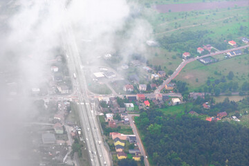 Naklejka premium Aerial view of small town partially covered by drifting clouds reveals roads, houses and green fields. Scenic perspective captures rural charm and contrast between urban layout and natural landscape
