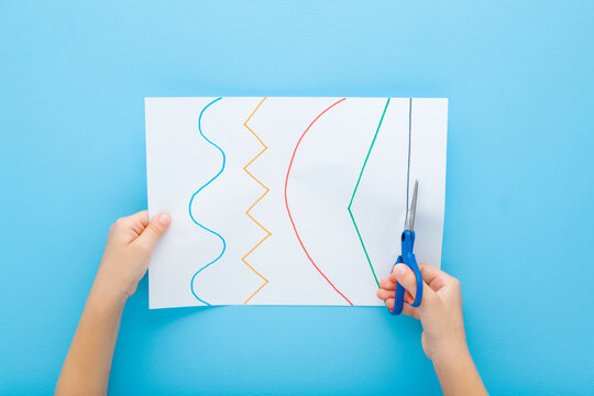 Little child boy hands holding scissors and cutting colorful different lines from white paper on light blue table background. Pastel color. Learning to use scissors. Point of view shot. Top down view.