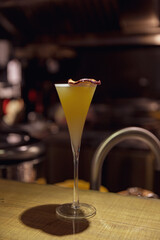 Yellow cocktail served in a tall stemmed flute glass, garnished with a dried citrus slice, set against a dark bar background with ambient lighting