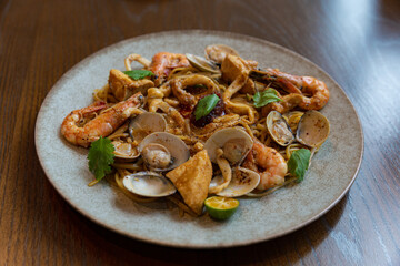 Asian-style spicy seafood pasta served with clams, shrimp, tofu, lime, and herbs on a rustic plate, showcasing a fusion of flavors and textures