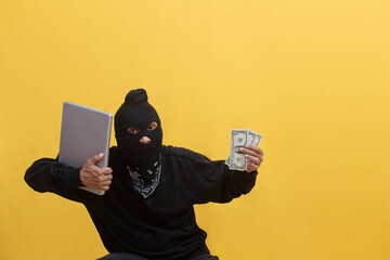 Portrait of a thief dressed in black wearing ski mask balaclava stealing laptop and dollar banknotes trying to escape after committing a crime. Ideal for theft, hacking, and crime concepts.