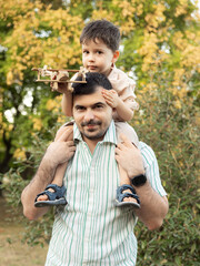 Happy family relaxing in park on summer day. Parents sit on blanket with smiling boy playing with wooden toy airplane. Father lifts child up, concept of flight, travel and joyful family moments.
