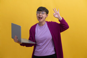 A cheerful young pretty woman celebrating success and feeling triumphant making ok sign gesture with hand while holding her laptop in the air showing both excitement and achievement.