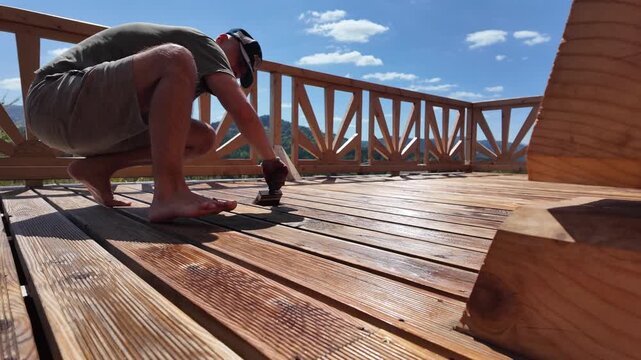 Man oils wooden deck boards with brush on a sunny summer day. Scenic mountain view, blue sky, and decorative railing highlight outdoor DIY wood protection and home improvement.
