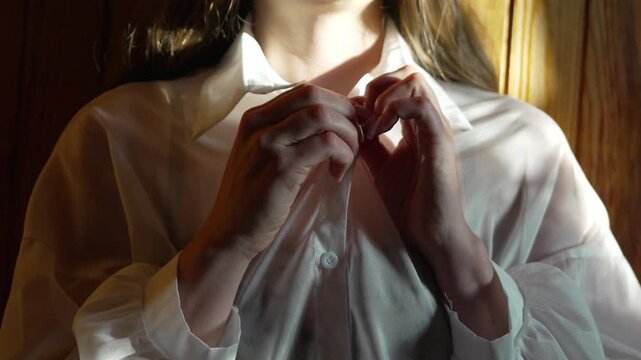 Woman unbuttoning shirt, close-up of hands unfastening buttons on white blouse in morning light
