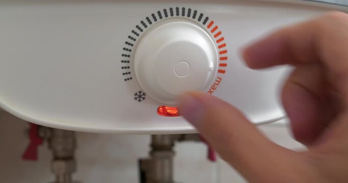 Regulator for heating cold water in boiler. Man turns temperature control on heater. Red light indicator comes on, boiler has turned on to heat water. Boiler heats water from cold water supply system
