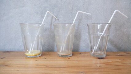 Three clear drinking glasses with white straws, mostly empty with drink remnants, placed on a wooden table against a concrete wall background.  
