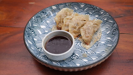 Crispy fried dumplings arranged on a patterned ceramic plate, accompanied by a small bowl of dark dipping sauce on a wooden table.  
