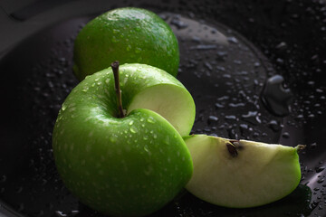 two vibrant green apples resting on glossy black surface, one apple sliced revealing its crisp interior, showcasing water droplets on their skin. close up.