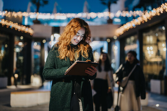A cheerful woman interacting with a tablet in a well-lit outdoor setting during nighttime. She appears happy and engaged, with festive string lights illuminating the area, creating a warm and vibrant