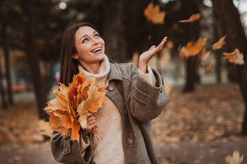 Young woman enjoying autumn park with colorful leaves in hand, portraying a cheerful and stylish...