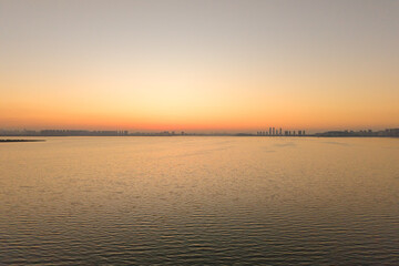 Fototapeta premium Sunset colors illuminate Tangxun Lake in Jiangxia District, Wuhan, China, as golden light reflects softly on the calm water, showing city buildings afar.