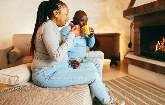 African american senior couple drinking hot chocolate in front of cozy fireplace at home - Winter lifestyle concept - Focus on woman face