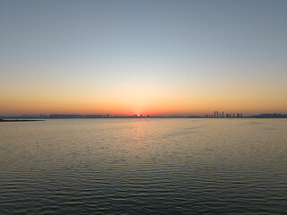 The warm light of a vibrant sunset spreads across the tranquil lake surface, as a modern city skyline appears on the distant horizon at dusk.