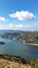 Obraz premium A serene vista of a lake nestled within a mountainous landscape, dotted with numerous wind turbines, under a vibrant blue sky filled with fluffy white clouds.