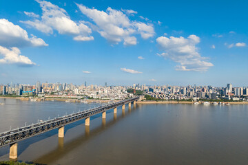 Obraz premium Wuhan China city skyline beautifully stretches along the Yangtze River, with a long bridge crossing its wide waters under a clear blue sky with white clouds.