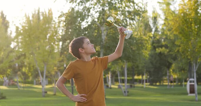 Happy cild boy holding trophy outdoors during golden hour. Kid boy celebrates his achievement victory smiling happily. Child boy feels proud energetic in summer park Child with cup in his hand sunset.
