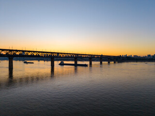 A grand bridge spans the vast Yangtze River in Wuhan, China, under a stunning sunset sky with barges moving peacefully on the water.
