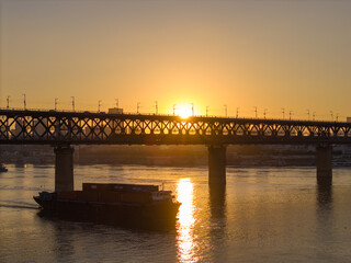 Fototapeta premium Golden sunset over the Yangtze River in Wuhan China shows a cargo ship moving under a large metal bridge with city skyline reflections.