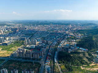 Obraz premium An expansive aerial view displays the sprawling urban panorama of Jiangxia, Wuhan. Numerous structures rise beside green hills, extending into the natural surroundings under a bright clear sky.