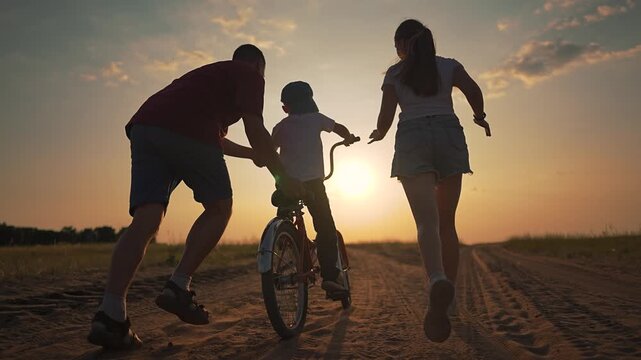 Boy rides red bike along dusty path while mother and father help from both sides during beautiful sunset. Parents support child learning to ride. Boy trains balance father and mother at sunset ride.
