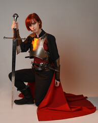 portrait of Fantasy warrior woman model, holding sword weapon, historical medieval armour costume with red fairytale cloak, strong fantasy knight. sitting kneeling pose isolated on studio background