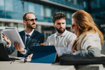 A group of three young professionals sitting together discussing work outdoors near offices, using a laptop and notebook.