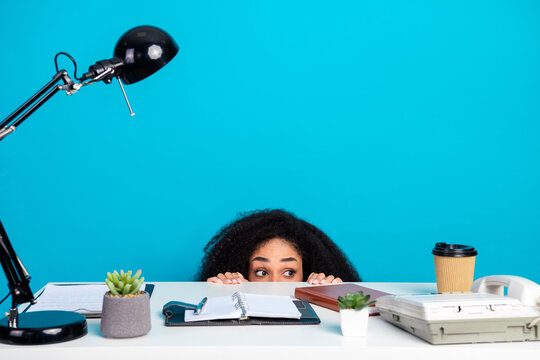 Playful young woman peeking over desk among office items, charming casual appearance against a solid blue background