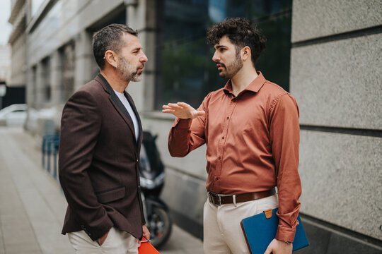 Two men having a discussion outside in an urban environment, one gesturing to emphasize a point while holding folders, representing communication and professional interaction. - Powered by Adobe
