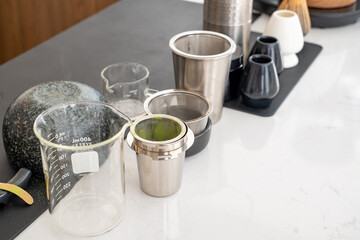 Close-up of tea making tools including stainless steel strainer, glass beaker, and ceramic cups on white countertop. Modern tea preparation equipment for brewing, serving, and beverage concept.
