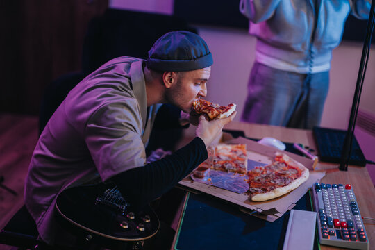 A man devours a slice of pizza in a home studio setting, surrounded by music equipment. The scene captures a relaxed and creative work atmosphere with focus on enjoying a casual meal. - Powered by Adobe