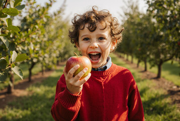 Joyful child in red sweater holding apple in orchard on sunny day, capturing autumn harvest excitement
