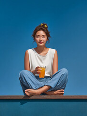 A young Korean woman sits on a brown wooden mat under a blue summer sky, holding a cool coral-colored drink with ice in her hand