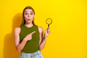 Adorable young woman poses with a magnifying glass on a vibrant yellow background, expressing surprise and charm in casual fashion